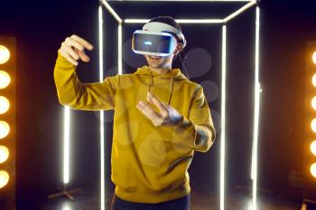 Young gamer plays simulator game in virtual reality headset and gamepad in luminous cube, front view. Dark playing club interior, spotlight on background, VR technology with 3D vision