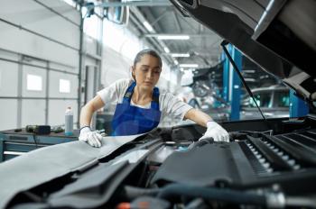 Female mechanic checks engine, car service. Vehicle repairing garage, woman in uniform, automobile station interior on background