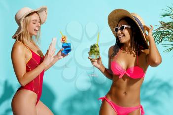 Smiling women in swimsuits poses with coctails on cyan background. Attractive girls in swimwear ready to take a tan. Models with slim body in swimming underwear, seductive female swimmers