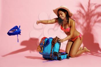 Happy woman in swimsuit unpacks a suitcase in studio, pink background. Girl in swimwear ready to take a tan. Model with slim body in swimming underwear, seductive female swimmer