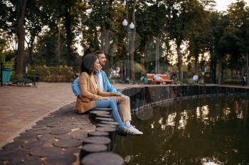 Love couple embracing at the pond in summer park. Man and woman relax outdoors, green lawn on background. Family hugging near the lake in summertime, weekend in nature