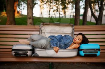 Young woman sleeping in opened suitcase on the bench in park. Female traveler with luggage leisures outdoors, passenger with bag resting in nature. Girl with baggage relax on city alley