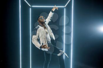 Stylish rapper in sunglasses poses in illuminated cube, dark background. Hip-hop performer, rap singer, break-dance performing, entertainment lifestyle