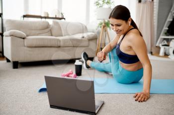 Smiling girl sits on the floor at home, online fit training at the laptop. Female person in sportswear, internet sport workout, room interior on background
