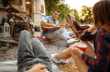 Cheerful friends sing songs with guitar on picnic at camping in the forest. Youth having summer adventure on rv, camping-car on background. Two couples leisures, travelling with trailer