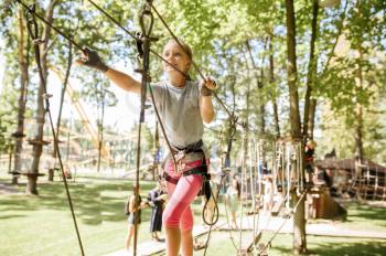 Little smiling girl in equipment climbs in rope park, playground. Child climbing on suspension bridge, extreme sport adventure on vacations, danger entertainment outdoors