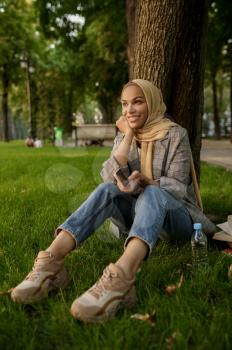 Smiling arab girl in hijab in summer park. Muslim woman resting on the lawn. Religion and education