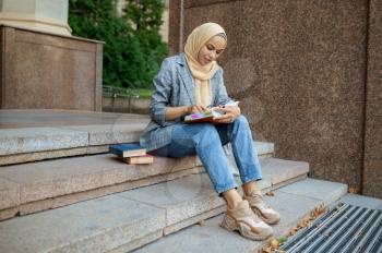 Arab female student sitting on the stairs in university. Muslim woman applies makeup at the entrance to the business center. Religion and education