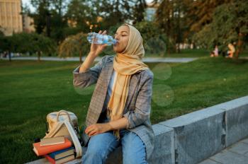 Arab female student in hijab drinks water in summer park. Muslim woman with books resting on the lawn. Religion and education