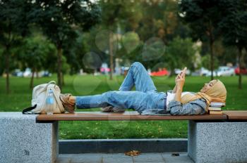 Arab female student lying on the bench in summer park. Muslim woman resting on the walking path. Religion and education