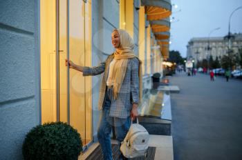Arab girl in hijab at the entrance of fashion store in downtown. Muslim woman walking on street. Female student in big city. Religion and education