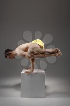 Yoga keeps balanc on hands in difficult pose on the pedestal, meditation position, grey background. Strong man doing yogi exercise, asana training, top concentration, healthy lifestyle