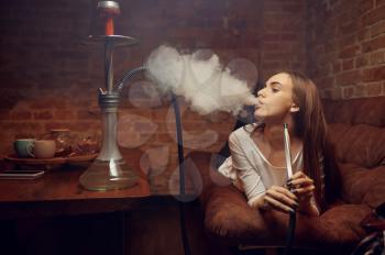 Young woman lying on sofa and smokes hookah in bar, chill out. Shisha smoking, traditional bong culture, tobacco aroma for relaxation, rest with hooka