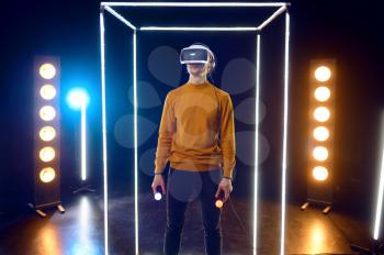 Gamer plays the game using virtual reality headset and gamepad in luminous cube. Dark playing club interior, spotlight on background, 3D VR technology