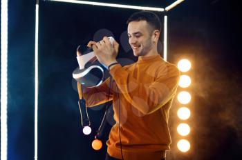 Young man holds virtual reality headset and gamepad in luminous cube. Dark playing club interior, spotlight on background, VR technology with 3D vision
