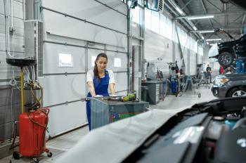 Female mechanic stands at the hood, car service. Vehicle repairing garage, woman in uniform, automobile station interior on background