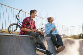 Two bmx bikers leisures in skatepark after training. Extreme bicycle sport, dangerous cycle exercise, street riding, biking in summer park