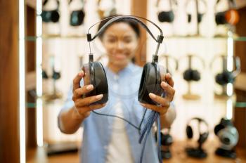 Woman shows vintage headphones in audio components store. Female person in music shop, showcase with earphones on background, buyer in multimedia salon