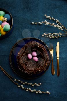 Blooming willow branch, easter eggs in nest and tableware on blue cloth background. Spring tree blossom and paschal food, fresh floral decoration for holiday celebration, event symbol