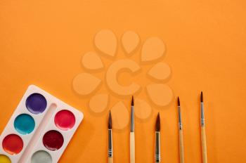 Watercolour paints and brushes closeup on orange background. Office stationery supplies, school or education accessories, writing and drawing tools