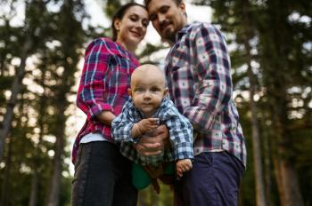 Mother, father and little baby poses in summer park. Mom and dad with male kid on lawn, picnic with child on plaid in the forest, family happiness