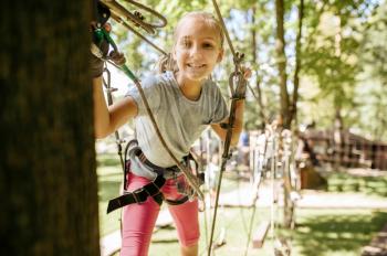 Little smiling girl in equipment climbs in rope park, playground. Child climbing on suspension bridge, extreme sport adventure on vacations, danger entertainment outdoors