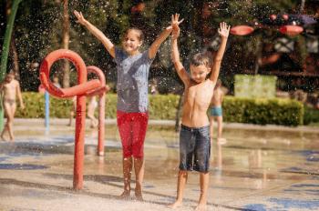 Little boy and girl play on water playground in summer park. Children leisures in aquapark, aquatic adventure on vacations, young brother and sister are having fun in fountain, waterpark