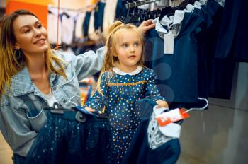 Mother and little baby choosing dress in kid's store. Mom and adorable girl near the showcase in children's shop, happy childhood, family makes a purchase in kid's market