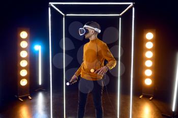 Gamer plays the game using virtual reality headset and gamepad in luminous cube. Dark playing club interior, spotlight on background, VR technology with 3D vision
