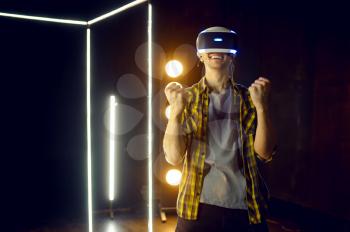 Happy man using virtual reality headset and gamepad in luminous cube, front view. Dark playing club interior, spotlight on background, VR technology with 3D vision
