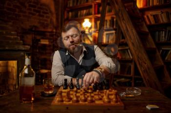 Man smokes a cigar, drinks alcohol beverage and play chess, bookshelf and vintage office interior on background. Tobacco smoking culture, specific flavor. Male smoker at the chessboard