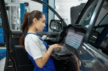 Female mechanic with laptop does the engine diagnostics, car service, professional inspection. Vehicle repairing garage, woman in uniform, automobile station interior on background
