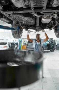 Male worker inspects car suspension, auto service. Vehicle repairing garage, man in uniform, automobile station interior on background