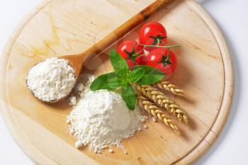 Pile of soft wheat flour, wooden spoon and fresh tomatoes on round cutting board