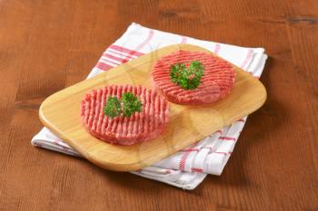 two raw hamburger patties with parsley on wooden cutting board and checkered dishtowel