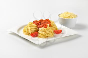 plate of cooked spaghetti with red pesto and bowl of grated parmesan cheese on white background