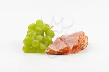 slices of ham and bunch of white grapes on white background