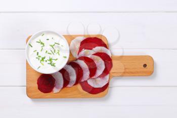 bowl of white yogurt with thin slices of beetroot and white radish on wooden cutting board