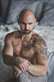 Bald beefy man with naked torso sitting in the room.
