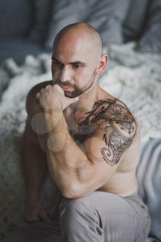 Bald beefy man with naked torso sitting in the room.