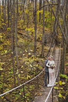 The bride and groom in beautiful outfits are on long wobbly suspension bridge.