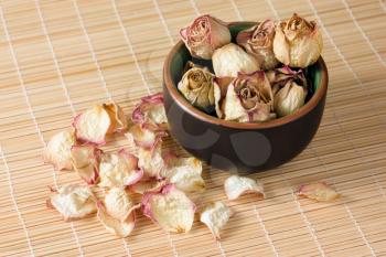 Dried rosebuds in the clay tea cup and dry rose petals on a straw mat closeup diagonal view