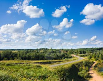 Road in countryside with trees and green grass around in sunny day under blue sky with white clouds