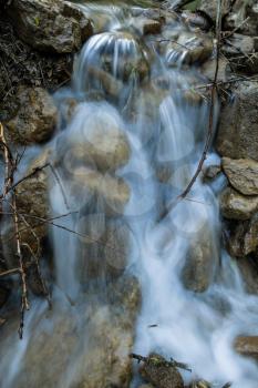 Small beautiful waterfall surrounded by rocks in river with stones  and small branches in forest in dusk closeup view. Natural summer landscape.