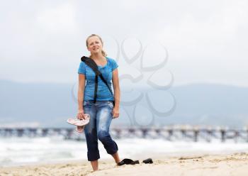 Smiling blonde woman with a backpack standing on the beach against the sea. Girl with backpack. Posing young woman. Selective focus on the model.
