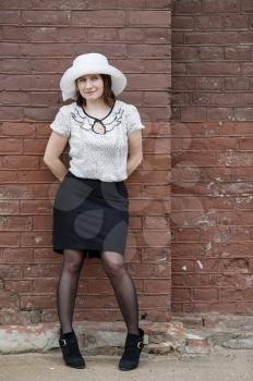 Pretty young fashion brunette woman in a white hat, blouse and black skirt, posing outdoor in old vintage brown brick wall background