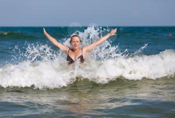 Smiling beautiful girl with her hands raised in the foam of a sea wave on a bright sunny day.