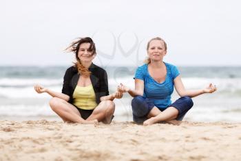 Girls relaxing outdoors.Two pretty young smiling women sitting in lotus pose on the sand on the beach and relax. Two girls resting on the beach. Selective focus on the models.