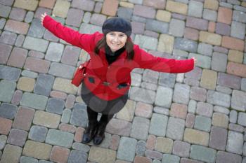 Smiling woman in a red jacket and a gray beret on a background of old paving slabs. Model posing, arms to the side and looking up at the camera. Top view. Shallow depth of field. Focus on the model's 