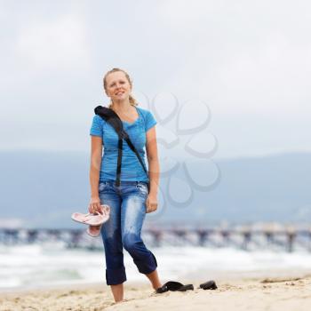 Young smiling blonde woman barefoot in blue t-shirt and jeans with a backpack standing on the beach against the sea.  Shallow depth of field. Focus on model.